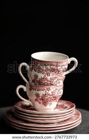 A creative, dramatic composition featuring a stack of vintage porcelain teacups and saucers with a classic red floral pattern, presented in an artistic layered or double-exposure style against a moody, dark background with high-contrast lighting.