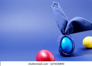 Creative easter bunny blue egg wrapped in a blue napkin and stands on a blue background. Nearby are yellow and red eggs. Minimalization trend blue monochrome, horizontal background