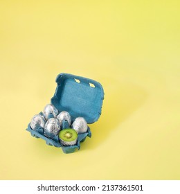 Creative Easter Concept Of Cardboard Box Of Eggs Painted In Silver Aluminum Foil And One Cut In Half And Inside Is Kiwi Fruit. Pastel Yellow Background.