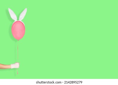 Creative easter minimal concept. Hand in white glove holding a pink balloon in bunny ears isolated on pastel green background. Easter Banner with copy space.
