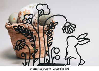 Creative Easter still life with a decorative line-art bunny sniffing a flower, standing in front of a rustic basket filled with colorful spring Easter eggs. Soft focus and bright background.