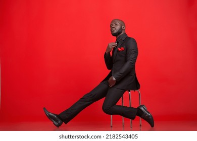 Creative emotional Portraits of a young mid-adult Kenyan black Male Man wearing a black complete suit black tie and red pocket size, black socks, and shoes against a red background in the studio