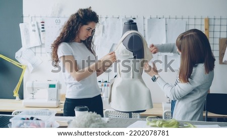 Creative fashion designers are pinning cut out pieces of fabric to mannequin while sewing women's garment in modern light studio. Ladies are concentrated on their work.