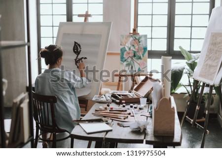 Creative female artist with paintbrush in her hair sitting in the art studio and drawing on the canvas while having art equipment on the table