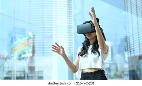 Creative Female Developer Wearing Virtual Reality Glasses, Interacting With Digital Interface While Standing Near In Large Window At Modern Office.