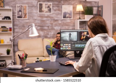 Creative female videographer working on a movie using her home computer. Boyfriend relaxing on sofa in the background. - Shutterstock ID 1585522510