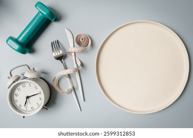 Creative flat lay composition with plate, alarm clock, knife, fork, dumbbell and measuring tape on gray background. Intermittent fasting, ketogenic, diet concept. top view, copy space.