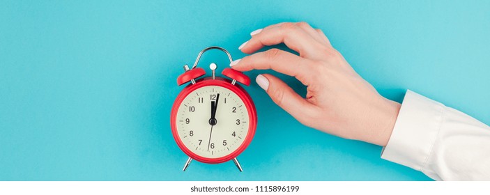 Creative flat lay concept top view of woman hand in white shirt holding the red vintage alarm clock on bright blue turquoise color paper background with copy space in minimal style, long wide banner