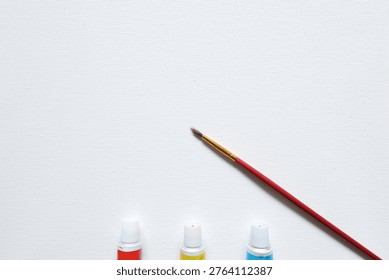Creative flat lay featuring diagonal paintbrush and primary color paint tubes on white textured surface with large copy space.