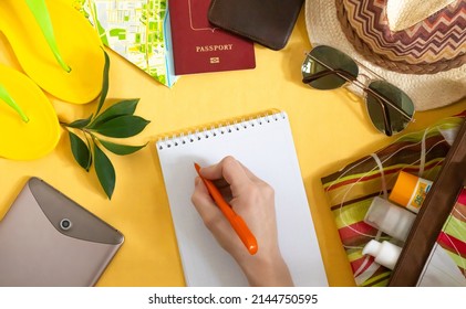 Creative flat lay of summer accessories supplies for travel preparation on yellow background. Flat lay travel with a female hand writing a list of things in a notebook.