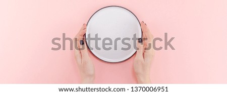Creative flat lay top view of woman hands black manicure with empty plate mock up on pastel millennial pink paper background copy space. Minimal dish service concept template for your text menu design