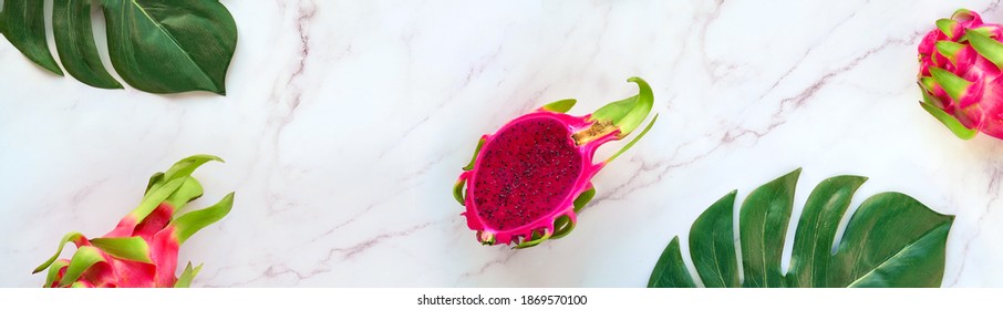 Creative flat layout with fresh organic pink dragon fruit, pitaya or pitahaya, on marble table with copy-space. Trendy top view, flat lay of dragonfruit with exotic monstera leaves.