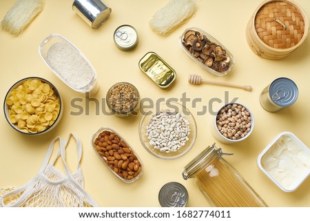 Creative flatlay with pantry staples. Glass jars with pasta, beans and chickpeas, canned goods, nuts and dried mushrooms in reusable containers. Top view pattern with basic products 