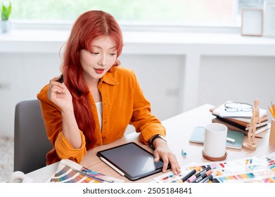 A creative and focused Asian female graphic designer with red hair is working at her desk in the studio, designing a new project and selecting colors from a set of swatches. - Powered by Shutterstock