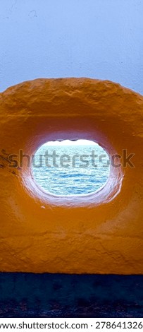 A creative frame-within-a-frame view of the blue ocean horizon, seen through an oval mooring chock or fairlead on a ship's bulwark. The textured, bright yellow painted steel creates a striking minimalist contrast against the blue water.