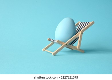 Creative funny composition with Easter egg while sitting on deck chair on pastel blue background. Minimal spring or summer vacation, holiday and travel concept.