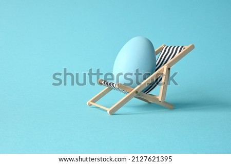 Creative funny composition with Easter egg while sitting on deck chair on pastel blue background. Minimal spring or summer vacation, holiday and travel concept.