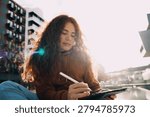 Creative ginger model using stylus to draw on digital tablet outdoors in sunset light. Talented artist creating digital illustration, graphic design sketching, online learning, remote freelance.