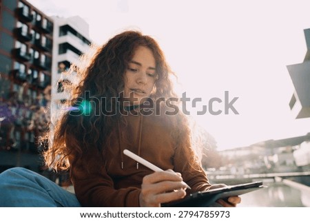 Creative ginger model using stylus to draw on digital tablet outdoors in sunset light. Talented artist creating digital illustration, graphic design sketching, online learning, remote freelance.