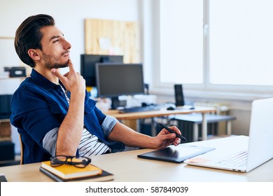 Creative Graphic Designer Looking For Inspiration. Young Man Sitting At Desk Thinking About New Ideas To Draw On Graphic Tablet.