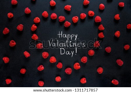 Creative greeting card Happy Valentine's Day. Frame of red raspberry berries in shape of heart with text inside Happy Valentine's Day written by chalk on black background. Top view. Flat lay.