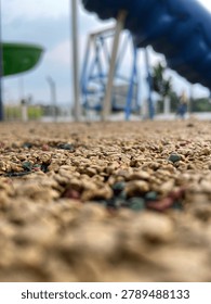 A creative ground-level perspective photograph capturing the textured playground surface with blurred playground equipment in the background. The shallow depth of field creates a dynamic composition that emphasizes texture, perspective, and outdoor recreational spaces.