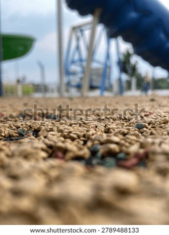 A creative ground-level perspective photograph capturing the textured playground surface with blurred playground equipment in the background. The shallow depth of field creates a dynamic composition that emphasizes texture, perspective, and outdoor recreational spaces.
