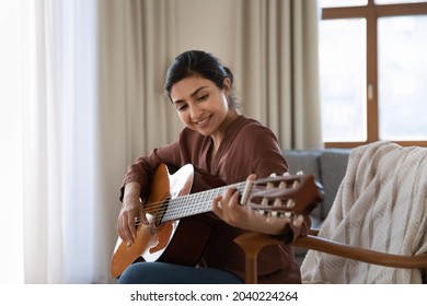 Creative hobby. Talented young mixed race female musician sit in armchair alone compose instrumental song using classic guitar. Smiling biracial lady play calm melody on musical instrument. Copy space