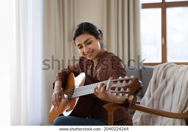 Creative hobby. Talented young mixed race female
musician sit in armchair alone compose instrumental song using
classic guitar. Smiling biracial lady play calm melody on musical
instrument. Copy space