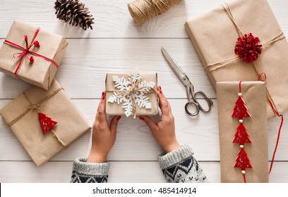 Creative Hobby. Woman's Hands Show Christmas Holiday Handmade Present In Craft Paper With Twine Ribbon. Making Bow At Xmas Gift Box, Decorated With Snowflake. Scissors On White Wooden Table, Top View.