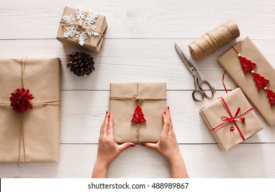 Creative Hobby. Woman's Hands Show Christmas Holiday Handmade Present In Craft Paper With Twine Ribbon. Scissors And Boxes With Bows And Snowflakes On White Wooden Table, Top View.