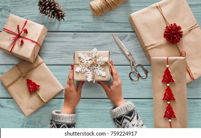 Creative Hobby. Woman's Hands Show Christmas Holiday Handmade Present In Craft Paper With Twine Ribbon. Making Bow At Xmas Gift Box, Decorated With Snowflake. Scissors On Blue Wooden Table, Top View.