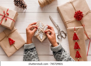 Creative Hobby. Woman's Hands Wrap Christmas Holiday Handmade Present In Craft Paper With Twine Ribbon. Making Bow At Xmas Gift Box, Decorated With Snowflake. Scissors On White Wooden Table, Top View.