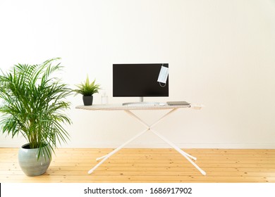 Creative Home Office Space During Coronavirus Quarantine, A Computer On Ironing Board With Medical Mask Hanging, Hand Sanitiser, Mouse, Keyboard And Plants