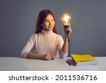 Creative idea, bright thinking, education, knowledge cognition. Portrait smart clever curious girl child with glowing lamp in hand looking at light bulb sit at desk isolated on grey studio background