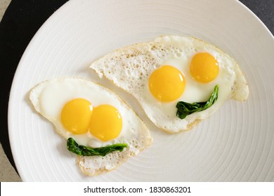 Creative idea of fried eggs in the form of funny ghosts. Halloween decor for kids' food. Feature - two eyes in one egg. White plate on a black background. Smiles made from spinach leaves.