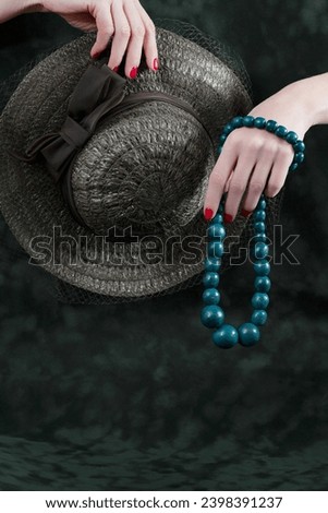 Creative image with hands holding woman old fashion vintage accessories hat with net and teal blue wooden beads neckless on a dark green background