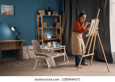 Creative latin female artist standing working new painting  Happy young lady painting canvas in her art studio  Cheerful young female artist working in her home workshop  copy space 