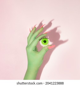 Creative layout with green painted hand with bright pink nails holding green eyeball against pastel pink background. Halloween celebration idea. Minimal flat lay concept.