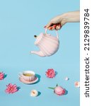 Creative layout with hand holding teapot and pouring tea into pink cup with with fresh flowers around on pastel blue background. Creative floral spring bloom concept. Natural herbal drink idea.