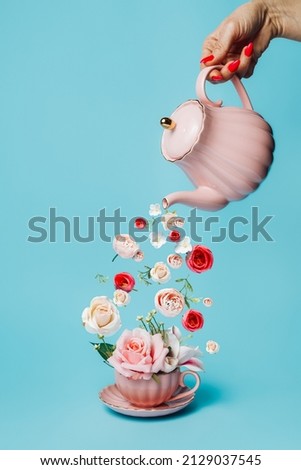 Creative layout with hand holding teapot and pouring fresh flowers and leaves into tea cup on pastel blue background. Creative floral spring bloom concept. Still life natural visual trend. 