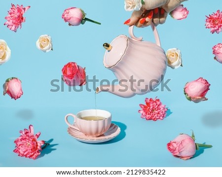Creative layout with hand holding teapot and pouring drink into tea cup with with fresh flowers flying on pastel blue background. Creative floral spring bloom concept. Still life natural visual trend.