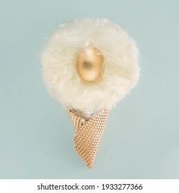 Creative Layout Made Of  Pastel White Fluffy Seed Hairs Nest With A Golden Egg In A Golden Ice Cream Cone. Minimal Flat Lay 2021 Easter Concept On Pastel Blue Background.