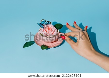 Creative layout with woman's hand holding teapot with fresh rose flower and butterfly on pastel blue background. Creative floral spring bloom concept. Minimal breakfast or drink still life idea.