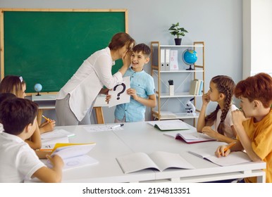 Creative Lesson. Friendly Female Teacher Whispers In Ear Of Little Schoolboy Giving Hint During School Lesson. Boy Is Holding Sign With Question Mark Standing In Front Of His Classmates In Classroom