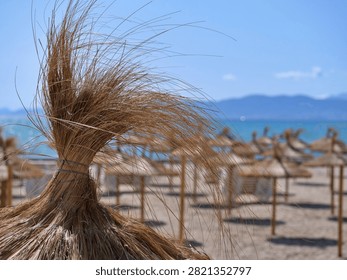 A creative low-angle macro perspective focusing on the dry, tied straw top of a traditional beach parasol. The background softly blurs out the sunny sandy beach, scattered umbrellas, turquoise sea, and blue horizon. Excellent for artistic travel layouts, summer holiday concepts, textures, natural craft materials, and peaceful tropical coastal aesthetics.
