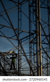 A creative low-angle shot looking through a dense web of scaffolding at a construction worker. The shallow depth of field emphasizes the geometric complexity of the metal pipes against a clear blue sky, highlighting the scale and detail of industrial labor.