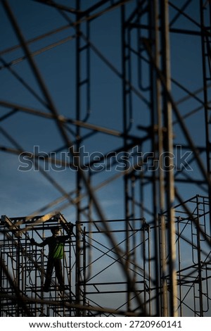 A creative low-angle shot looking through a dense web of scaffolding at a construction worker. The shallow depth of field emphasizes the geometric complexity of the metal pipes against a clear blue sky, highlighting the scale and detail of industrial labor.