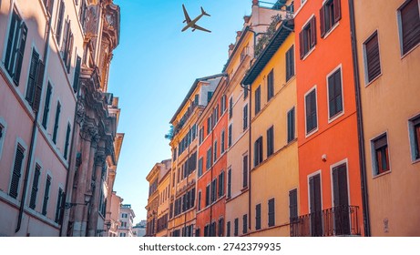A creative low-angle view of an airplane in a clear blue sky, flying between tall, colorful buildings in a classic Italian style. Captured in the historic heart of Rome, near Trastevere or Monti, this shot highlights the contrast between centuries-old architecture and modern technology