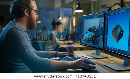 Creative Male Game Developer Works with Graphics for the New Level Design, He Sits at His Wooden Table Working on Two Display Personal Computer. Other People Work in This Creative Studio.